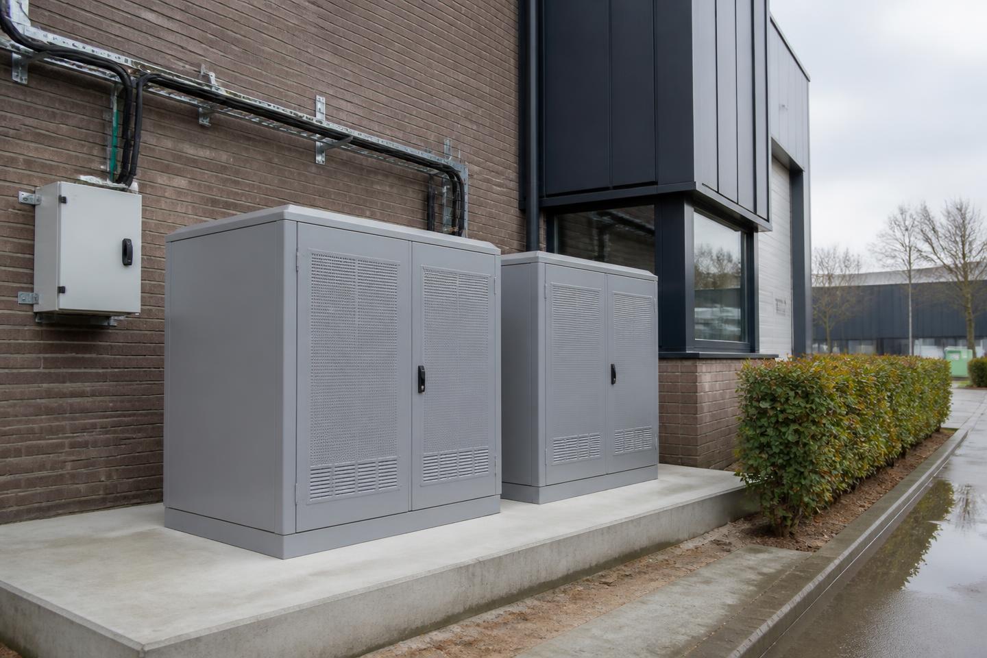 Twee grijze batterijkasten op een betonnen fundering naast een Nederlands bedrijfspand